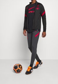 Sort Nike zip-jakke med røde accenter, sort Liverpool t-shirt, grå tætsiddende bukser med røde detaljer og sorte fodboldstøvler med orange snørebånd. Fodbold i nærheden.