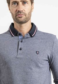 Polo gris avec un motif texturé, col marine présentant des rayures rouges et blanches, patte de boutonnage à trois boutons et un petit logo sur la poitrine.