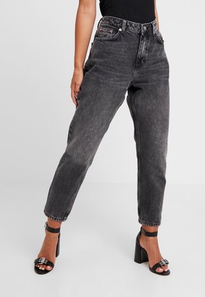 Persoon die hoge taille vervaagde zwarte jeans en zwarte sandalen met hak en enkelbandjes draagt, staand tegen een effen grijze achtergrond.