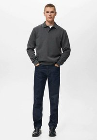 Dunkelgrauer Kragenpullover, kombiniert mit dunkelblauen Jeans. Das Model steht mit den Händen in den Taschen und trägt schwarze Schuhe. Einfarbiger Hintergrund.