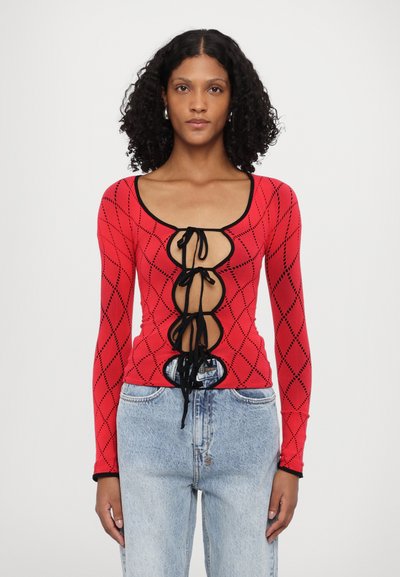 Haut rouge tricoté à manches longues avec un motif en losange, présentant un décolleté arrondi, des accents noirs et des détails de laçage à l'avant. Porté avec un jean bleu clair.