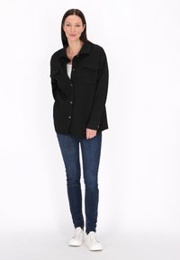Jachetă căptușită oversized neagră, cu închidere prin butoane, două buzunare pe piept și guler, purtată cu blugi albaștri și teniși albi.