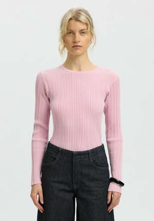 Jeune femme aux cheveux blonds portant un haut à manches longues côtelé rose clair et un jean foncé taille haute sur un fond uni.