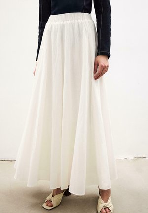 Γυναίκα που φοράει μια ροή λευκή μακριά φούστα, μαύρο μακρυμάνικο τοπ και μπεζ στριφτά ανοιχτά σανδάλια με τακούνι, στέκεται σε ουδέτερο πάτωμα.