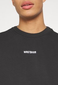 Personne portant un t-shirt noir côtelé à col rond avec le texte brodé en blanc "WRSTBHVR" centré sur la poitrine.