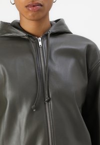 Felpa in pelle grigia con zip frontale, dettagli con cordoncino e texture liscia. Presenta un cappuccio e accenti in metallo.