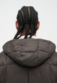 Pufferjakke i mørkegrå med quiltet tekstur og hette, med flettede hår som er synlige bak.
