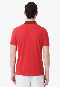 Röd polo skjorta med korta ärmar, brun krage och subtilt logotypdetalj på baksidan. Tillverkad av en mjuk tyg med en figurnära design.