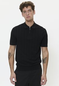 Black short-sleeve polo shirt with a textured pattern, zip collar, and ribbed hem. The model has a tattoo on his forearm.