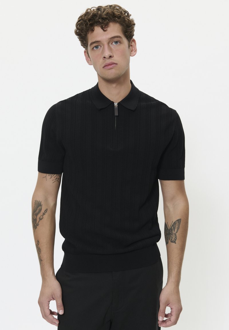 Black short-sleeve polo shirt with a textured pattern, zip collar, and ribbed hem. The model has a tattoo on his forearm.