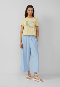 Lichtgele t-shirt met "Aloha" graphic, gecombineerd met losse lichtblauwe broek en witte sandalen met bandjes. Plat, relaxte pasvorm, zachte stof.