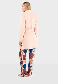 Robe lunga a maniche lunghe di colore rosa chiaro con una cintura; abbinata a pantaloni a fantasia con fiori blu e rossi su uno sfondo chiaro.