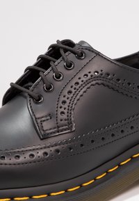 Chaussure de ville en cuir noir avec un bout arrondi, perforations brogue, lacets gris et surpiqûre jaune sur la semelle. Texture lisse et finition brillante.