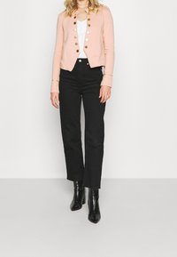 Veste rose pâle avec boutons dorés, coupe courte et manches longues. Associée à un pantalon noir à jambes larges et des bottines pointues noires.