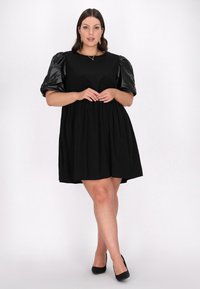 Rochie neagră cu un corset ajustat și o fustă evazată. Prezentă manșete lucioase și bombate și un decolteu rotund. Stilizată cu tocuri negre.