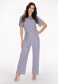 Grijze jumpsuit met korte mouwen, een ceintuur in de taille en wijde, vloeiende benen. Gladde stof met een subtiele glans, een paar zilveren sandalen met bandjes.