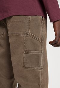 Bruna cargobyxor har flera fickor, ett texturerat tyg och förstärkta sömmar. Matchas med en vinröd långärmad tröja.