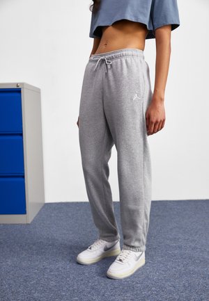 Vrouw draagt grijze joggingbroek met trekkoord en witte sneakers, staand op een blauw tapijt vlakbij een blauwe archiefkast.