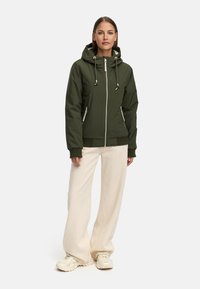 Grön zip-jacka med huva, elastiska mudd och framfickor. Buren tillsammans med krämfärgade vida byxor och ljusa sneakers.