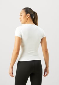 Top blanco de manga corta ajustado, hecho de un tejido suave, combinado con leggins negros ajustados; visto desde atrás.