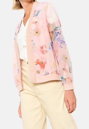 Femme portant une veste légère transparente à fleurs rose clair par-dessus un haut blanc et un pantalon couleur crème, debout devant un fond uni.