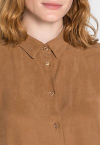 Hnědá košile s límečkem z texturované látky, vybavená kulatými hnědými knoflíky a hladkým povrchem. Detailní pohled na knoflíkovou légu.