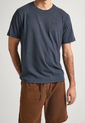 Mand iført en ensfarvet mørkegrå t-shirt med korte ærmer og brune shorts med snøre, stående mod en ensfarvet lys baggrund.