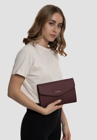 Clutch in pelle bordeaux con finitura testurizzata, dotato di chiusura a pattina e logo impresso. Il modello tiene la borsa contro uno sfondo neutro.