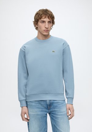 Junger Mann trägt ein hellblaues Sweatshirt mit langen Ärmeln und blaue Jeans, steht mit neutralem Gesichtsausdruck vor einem schlichten Hintergrund.