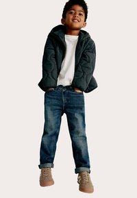 Donkere gewatteerde jas, wit T-shirt, blauwe spijkerbroek, beige sneakers. De jas heeft een capuchon en rits; de jeans zijn iets omgeslagen bij de boorden.