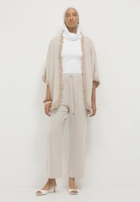 Frau mit weißem Kopftuch, weißem Oberteil, beigem Fransenstola, lockeren beigen Hosen, weißen offenen Schuhen und Holzarmbändern, die vor einer schlichten weißen Wand steht.