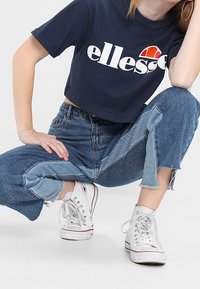 Mörkblå kortärmad t-shirt med vitt "ellesse"-logotyp. Högmidjade, slitna, i två nyanser av blå jeans. Vita högklackade sneakers med röda detaljer.