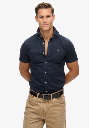 Camisa de manga corta azul marino con collar tipo button-down, bolsillo frontal y botones claros, combinada con pantalones beige y un cinturón de cuero negro.