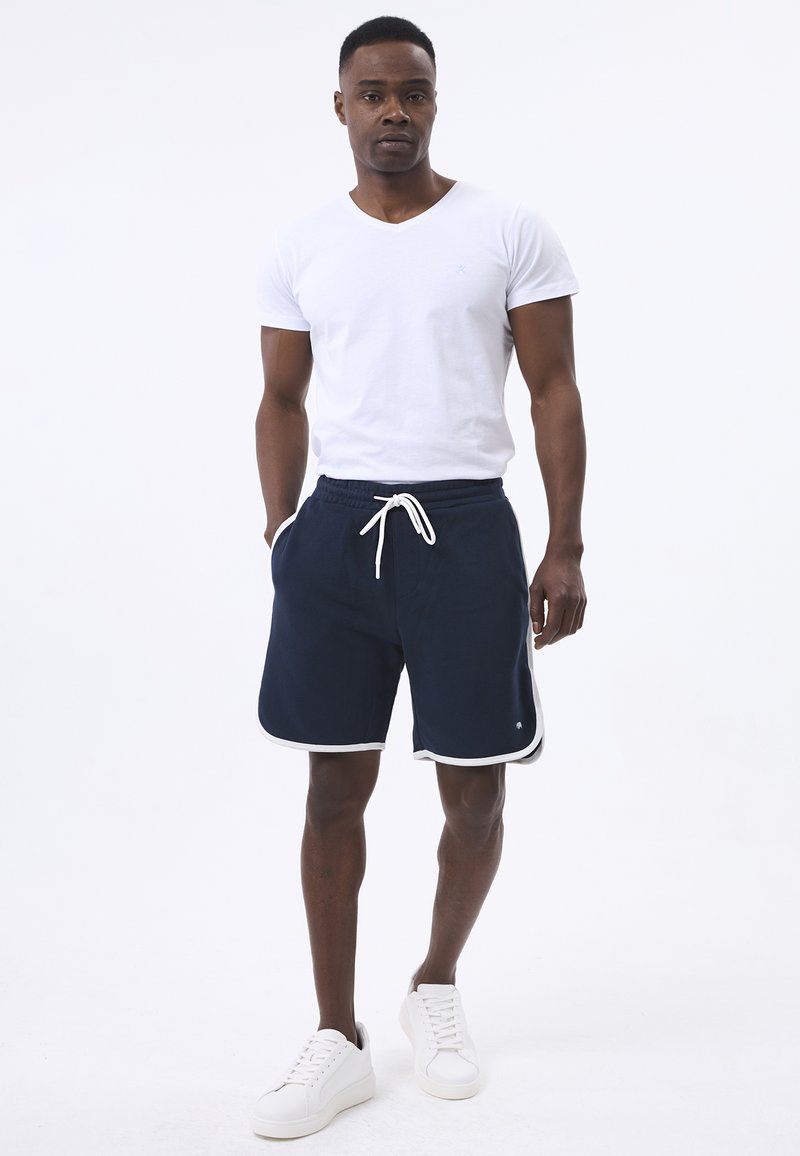 Navyblauwe sportshorts met witte zijstrip en trekkoord in de taille, gecombineerd met een eenvoudige witte V-hals t-shirt en witte sneakers.