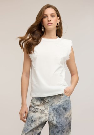 Jeune femme aux longs cheveux bruns portant un haut blanc sans manches aux bords effilochés et un pantalon large à motif floral dans des tons gris.