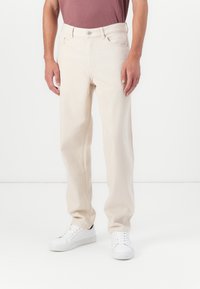 Pantalones de mezclilla beige con un corte relajado, que cuentan con un diseño de cinco bolsillos y un cierre de botón. Combinados con zapatillas blancas.