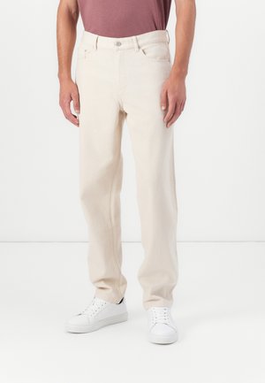 Pantalons en denim beige à coupe décontractée, présentant un design à cinq poches et une fermeture à boutons. Associés à des baskets blanches.