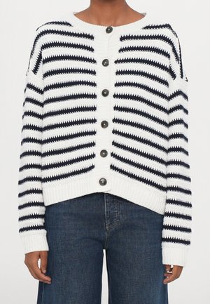 Cardigan tricoté blanc à rayures horizontales noires et boutons noirs, porté avec un jean en denim bleu foncé.