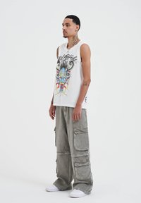 White sleeveless shirt with a colorful graphic design featuring a skull and horns, paired with loose grey cargo pants and white sneakers.