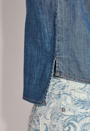 Spijkerblouse met een donkerblauwe textuur en oranje stiksel, voorzien van een zijsplit. Gecombineerd met lichtblauwe broek met patroon.