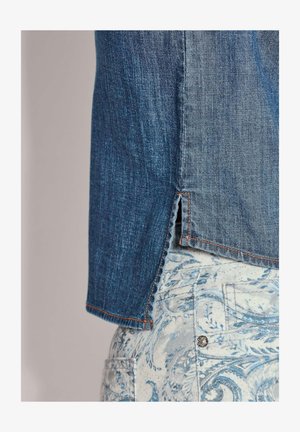 Spijkerblouse met een donkerblauwe textuur en oranje stiksel, voorzien van een zijsplit. Gecombineerd met lichtblauwe broek met patroon.