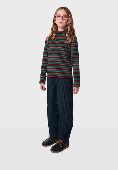 Vihreä-punaraidallinen pitkähihaisen paita yhdistettynä tummiin liituraitahousuihin. Jalkineet ovat mustat, joissa on ruskeat nauhat ja yksityiskohdat.