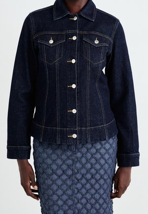 Kvinde iført en mørkeblå denimjakke med knaplukning og et blåt tekstureret skørt, stående mod en ensartet lys baggrund.