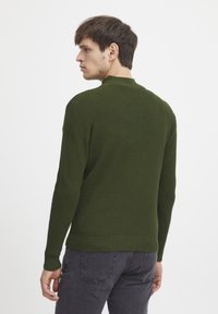 Maglione verde lavorato a maglia con superficie testurizzata, caratterizzato da un colletto alto e maniche lunghe, abbinato a jeans grigio scuro.