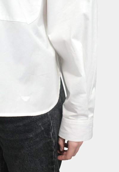 Camicia bianca a maniche lunghe con una consistenza liscia, dotata di un orlo curvo e di un piccolo logo sul retro. Abbinata a jeans in denim scuro.