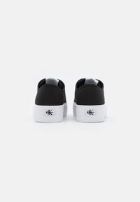 Svarta canvas sneakers med vita gummisulor, med en minimalistisk design och en logotypdetalj på hälen. Bakifrån.