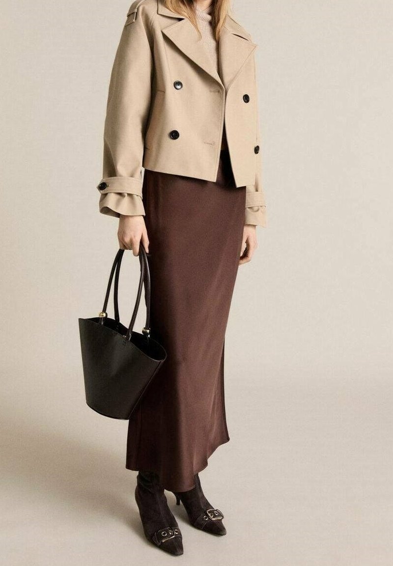 Veste beige à double boutonnage, jupe maxi en satin marron, sac à main noir et bottes à talons noires avec des accents de boucle.