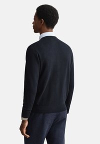 Juodas megztas megztinis su gofruotais rankogaliais ir apačia, dėvimas ant balto marškinio su apykakle. Fonas vientisa balta spalva, akcentuojant drabužio detales.