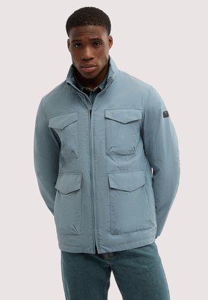 Mand iført lyseblå lynlåstrøje med fire klaplommer og blå jeans, stående mod en ensartet lys baggrund.