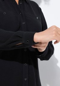 Schwarzes Hemd mit Knöpfen aus strukturiertem Stoff, mit Kragen, zwei Brusttaschen und kreisförmigen Hardware-Details an den Knöpfen und Manschetten.